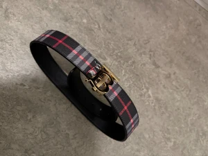 Burberry rutig skinnbälte med guldspänne - Säljer ett snyggt Burberry bälte i svart skinn med klassiskt rutig mönster i grått och rött. Bältet har ett stilrent guldspänne med Burberrys ikoniska B-logga och detaljerade vita emblem längs bandet. Perfekt för att lyfta din outfit med en lyxig touch.