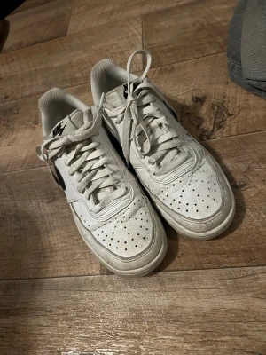 Nike Air Force 1 vita sneakers - Nike Air Force 1 sneakers i vitt skinn med svart swoosh på sidan. Klassisk låg modell med rund tå, snörning och perforerade detaljer på tån för extra ventilation. Snygg och tidlös design som passar till de flesta outfits.