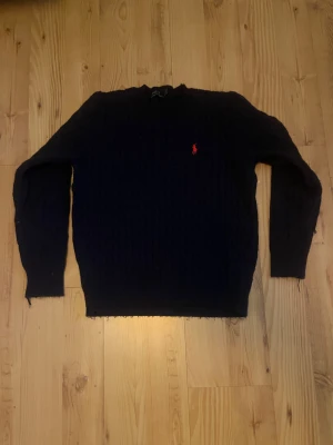 Mörkblå stickad tröja Polo Ralph Lauren - Mörkblå stickad tröja från Polo Ralph Lauren i storlek M. Klassisk kabelstickad design med röd broderad logga på bröstet. Rund hals och långa ärmar med ribbade muddar. Perfekt för dig som gillar stilrena och tidlösa plagg. Har några lösa trådar som syns på bilderna men trådarna märks knappt av när tröjan är på. Priset är inte hugget i sten