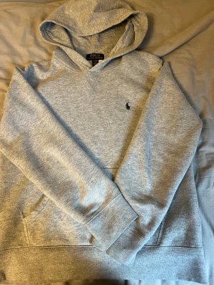 Grå hoodie från Polo Ralph Lauren - Snygg grå hoodie från Polo Ralph Lauren med klassisk broderad logga på bröstet. Tröjan har huva, långärmade ärmar och en stor magficka. Mjuk och skön bomullsmix som passar perfekt när du vill vara chill och ändå se fräsch ut. Storleken är L/14-16. Var inte rädda att fråga om något ni undrar om, priset kan sänkas!!