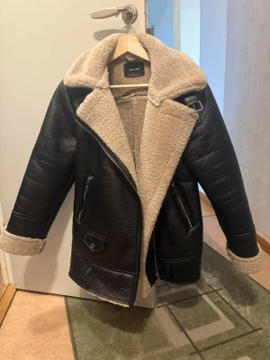 Svart teddyjacka från Stradivarius - Säljer en svart teddyjacka från Stradivarius i storlek S. Jackan har mjukt beige foder, bred krage, dragkedja och detaljer med spänne vid halsen. Utsidan är i konstläder-look och insidan är fluffig teddy. Perfekt för kalla dagar och riktigt snygg till jeans.