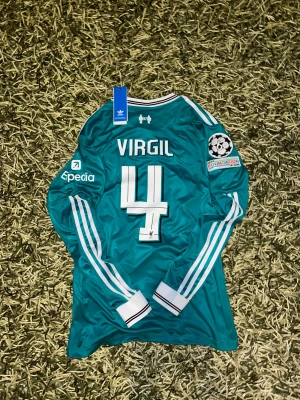 Liverpool Virgil #4 långärmad tröja L - Liverpool långärmad fotbollströja i turkosblå färg med vita detaljer. Broderat klubbmärke, tryck med Virgil och nummer 4 på ryggen, Champions League-märke på ärmen och klassiska Adidas-ränder. Materialet är glansigt och lätt, perfekt för match eller träning.