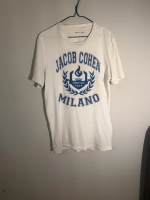 Vit t-shirt Jacob Cohen Milano - Vit t-shirt från Jacob Cohen med blått tryck framtill där det står 'Jacob Cohen Milano' och en grafisk logga. Klassisk rund halsringning och korta ärmar. Tillverkad i 100% bomull för en skön och mjuk känsla. Finns en otroligt liten fläck på magen som knappt syns och speciellt inte vid användning.