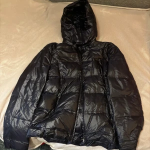Tunn pufferjacka med huva - Säljer en svart/mörkblå pufferjacka med huva och dragkedja framtill. Jackan har en quiltad design och är tillverkad i glansigt syntetmaterial. Den är väldigt tunn och liten i passform. Inget jag rekommenderar för vintern utan perfekt för våren och hösten. Inga prisförslag!