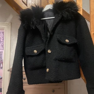 Svart teddyjacka med fuskpäls - Säljer en svart teddyjacka med fluffig fuskpäls runt kragen och stora fickor med knappar framtill. Jackan är croppad i modellen. Perfekt för dig som vill ha en mysig och trendig look i höst och vinter. Passar xs-m beroende på hur man vill att den ska sitta. 