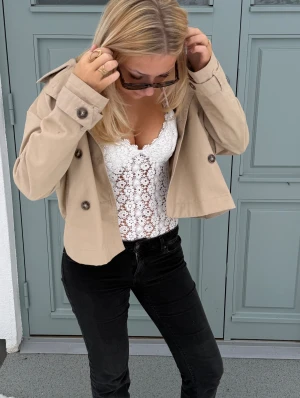 Vit spetsbody  - Supersöt vit spetsbody med tunna axelband och blommigt spetsmönster. Toppen är tight och har en feminin v-ringad halsringning med fina spetsdetaljer längs kanterna. Perfekt att styla med jeans eller kjol för en fräsch look.