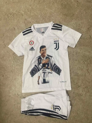 Juventus Ronaldo fotbollströja set - Juventus fotbollströja och shorts i vitt med svarta detaljer. Tröjan har tryck av Cristiano Ronaldo, nummer 7 och Juventus-logga. Adidas-märke och Climalite-material. Svarta ränder på axlar och shorts, samt stora tryck på både fram- och baksida.
