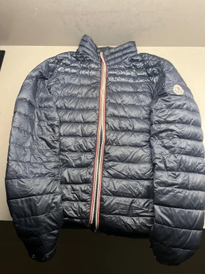Mörkblå Moncler pufferjacka herr - Snygg mörkblå pufferjacka från Moncler med klassisk logga på ärmen och coolt randigt band längs dragkedjan. Jackan har quiltad design, lång ärm och stårkrage. Perfekt för dig som vill ha en stilren och varm jacka med streetvibe.
