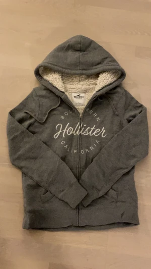 Grå hoodie med teddyfoder Hollister - Mysig grå hoodie från Hollister med fluffigt vitt teddyfoder på insidan. Tröjan har huva, dragkedja framtill och tryckt logga på bröstet. Perfekt för kalla dagar och chill hemma. Snygg och enkel att matcha med jeans eller mjukisbyxor.