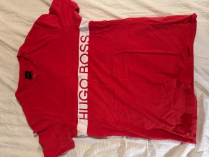 Röd Hugo Boss t-shirt med logga - Röd t-shirt från Hugo Boss med vit logga tryckt över bröstet. Modellen är slim fit med korta ärmar och rund halsringning. Tillverkad i mjuk bomull som känns skön mot huden. Perfekt för dig som gillar stilrena och sportiga plagg.