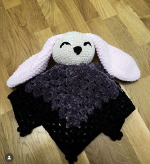 Kanin snuttefilt - Bilderna visar en handgjord virkad snuttefilt med ett gulligt kaninhuvud och långa rosa öron. Filten är mjuk och har en lila och svart färgskala, perfekt som present till någon som gillar hantverk eller vill ha en mysig accessoar. Passar dig som gillar unika, kreativa och personliga saker.