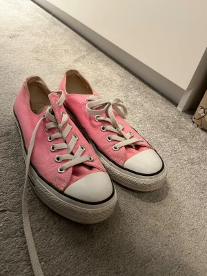 Rosa Converse All Star sneakers - Säljer ett par klassiska Converse All Star sneakers i en snygg rosa färg med vita snören och tåhätta. Skorna har lågt skaft, rund tå och är tillverkade i textil med gummisula. Perfekta för dig som vill ha en färgklick till outfiten.