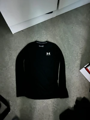 Svart långärmad träningströja Under Armour - Två tränings tröjor. Svart långärmad träningströja från Under Armour med vit logga på bröstet. Tillverkad i ett syntetmaterial som andas och har kompressionseffekt, perfekt för intensiva träningspass. Snygg och stilren design med rund hals.