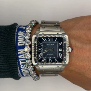 Seiko automatisk fyrkantig klocka silver - Stilren Seiko automatisk klocka med fyrkantigt boett i borstat silverfärgat stål och matchande länkarmband. Urtavlan är mörkblå med romerska siffror och datumvisning. Snygg detalj med synlig mekanism på baksidan. Perfekt för dig som gillar klassisk men modern stil.