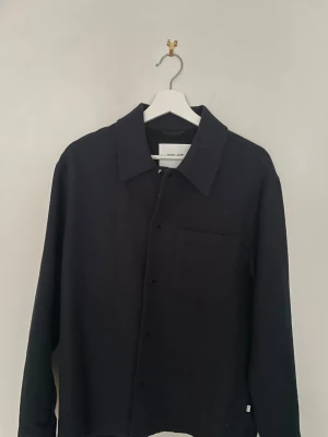 Svart tröja från NN.07 i ull - Stilren svart tröja från NN.07 med klassisk krage och bröstficka. Materialet är mjuk ull, designat av Beste i Italien. Skjortan har knappar framtill och lång ärm, perfekt för dig som gillar minimalistisk och clean look.