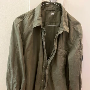 Olivgrön overshirt från C.P. Company - Säljer en olivgrön overshirt från C.P. Company med klassisk krage, dragkedja och knappar framtill. Skjortan har lång ärm, ficka på ärmen med den ikoniska C.P. Company-linsen och är tillverkad i bomull. Perfekt lager-på-lager-plagg med snygg streetkänsla.