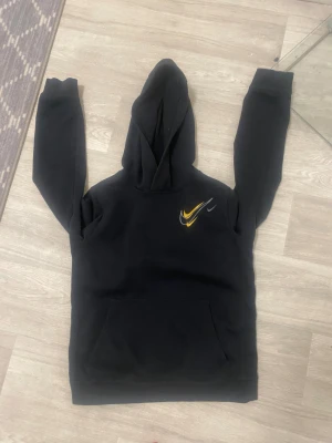 Svart hoodie från Nike med gul detalj - Svart hoodie från Nike i storlek xs med klassisk huva och magficka. På bröstet finns två Nike-loggor i gul och grå färg som ger en snygg kontrast. Tröjan har långa ärmar och är tillverkad i mjuk bomull, perfekt för chill dagar. Den är bra skick inga defekter eller hål. Passar för dig som är 160-170cm lång. Om du undrar nåt hör av mig. Priset går att diskuteras.