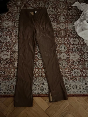 Bruna skinnbyxor med slits - Snygga bruna skinnbyxor med hög midja och raka ben. Byxorna har en stilren look med slits vid bensluten som ger en extra edge. Perfekta för dig som vill sticka ut med en cool vibe. Materialet är mjukt och följsamt syntetskinn.