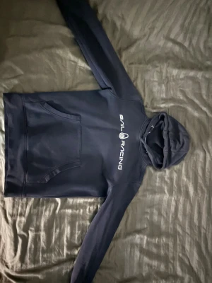 Mörkblå Sail Racing hoodie (PASSAR ÄVEN XS) - Snygg mörkblå hoodie från Sail Racing med vit logga tryckt på bröstet. Tröjan har huva med snörning och en stor magficka framtill. Perfekt för dig som gillar sportig och avslappnad stil. Långärmad och tillverkad i mjukt material för extra komfort.