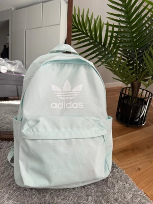 Ljusblå Adidas ryggsäck - Snygg ljusblå ryggsäck från Adidas med klassisk logga framtill. Väskan har ett stort huvudfack, en framficka med dragkedja och vadderade axelremmar med Adidas-tryck. Perfekt storlek för skola eller häng. Materialet är slitstark polyester.