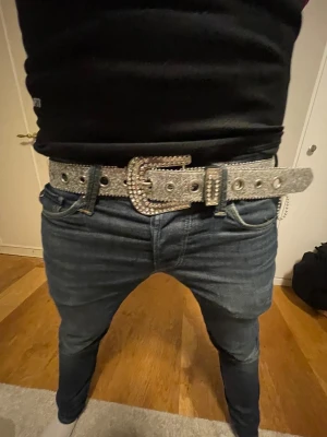 Glittrigt silverbälte - Säljer ett brett bälte i silver med glittrig yta och stora nitar längs hela bandet. Spännet är dekorerat med stenar och ger en riktigt iögonfallande look. Perfekt för dig som vill sticka ut och addera extra bling till outfiten.