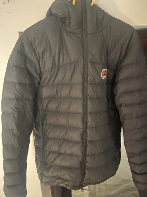 Svart Fjällräven Expedition dunjacka M - Säljer en svart Fjällräven Expedition Series dunjacka i storlek M. Jackan har puffad design, huva med gul snörning och klassisk Fjällräven-logga på bröstet. Perfekt för kalla vinterdagar med sin värmande dunfyllning och praktiska dragkedja framtill.