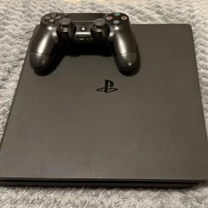 PlayStation 4 Slim - Sony PlayStation 4 Slim med en original DualShock 4-kontroll och tillhörande kablar. Konsolen och kontrollen är i gott skick med normalt slitage. En av laddningskablarna har tejp vid kontakten eftersom den har slitits men funkar bra annars. Perfekt för spel och underhållning. Kan diskutera pris!