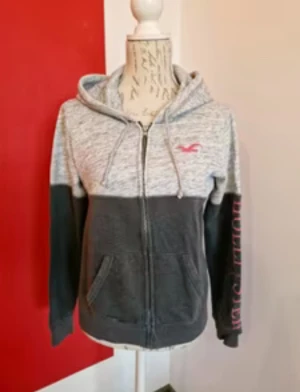 Grå och mörkgrå kofta Hollister - Snygg tvåfärgad hoodie från Hollister med ljusgrå ovandel och mörkgrå nederdel. Tröjan har dragkedja, huva med snörning och fickor framtill. Rosa Hollister-logga på bröstet och tryck på ena ärmen. 