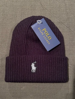 Svart ribbad mössa Polo Ralph Lauren - Snygg svart ribbad mössa från Polo Ralph Lauren med klassisk broderad logga framtill. Mössan är stickad och har en uppvikt kant för extra stilpoäng. Perfekt accessoar för att hålla dig varm och se cool ut under kyliga dagar. På bilden ser mössan mer lila ut men är helt svart