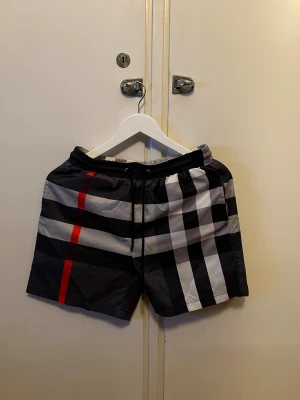 Burberry bad shorts  - Säljer ett par sjukt snygga Burberry badshorts i storlek M. De har klassiskt rutigt mönster i svart, vit och grå med en fet röd rand. Shortsen har elastisk midja med snörning och meshfoder på insidan. Perfekt för sommaren och strandhäng!
