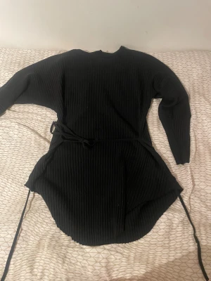 Svart ribbad tröja från Ezgi - Svart ribbad tröja från Ezgi i medium, med lång ärm och rund hals. Tröjan har ett knytband i midjan på både insidan och utsidan för snygg passform och är tillverkad i ett stickat material som känns mjukt och bekvämt. Perfekt för dig som gillar stilrena och enkla plagg.