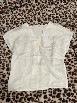 Vit blus med v-ringning från Vila - Säljer en stilren vit blus från Vila i storlek 36. Blusen har en snygg v-ringning, korta ärmar och är gjord i ett mjukt, slätt material som ger en skön känsla. Perfekt att matcha med jeans eller kjol för en clean look.