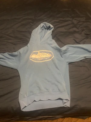 Blå hoodie från Corteiz med tryck - Säljer en blå hoodie från Corteiz med ett stort vitt tryck framtill som föreställer ett skepp. Hoodien har huva, magficka och ribbade muddar. Perfekt för dig som gillar streetwear och vill sticka ut med en unik design.