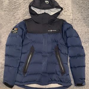 Sail Racing Drift Jacket W - Säljer en blå och svart pufferjacka från Sail Racing med huva och coola detaljer som patch på ärmen och logga på bröstet. Jackan har två snedställda fickor med dragkedja och justerbara muddar. Perfekt för kalla dagar när du vill vara både varm och snygg. Storlek L, men passar M/S.😊