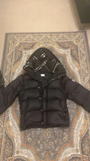 Svart Burberry pufferjacka med huva - Svart pufferjacka från Burberry med avtagbara ärmar, vilket gör att den kan användas som väst. Jackan har en stor huva med klassiskt rutigt Burberry-foder i svart, grått och vitt. Två dragkedjefickor framtill och tryckknappar. Snyggt Burberry-tryck på ena ärmen.