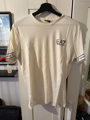 Vit EA7 Emporio Armani t-shirt - Snygg vit t-shirt från EA7 Emporio Armani med svart logga på bröstet och tryck på ärmarna. Klassisk rund hals och korta ärmar. Tillverkad i mjuk bomull som känns skön mot huden. Perfekt för dig som gillar stilrena och exklusiva plagg.