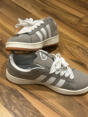 Adidas Campus - Skorna har breda vita snören och en platt sula i vitt gummi. Ovandelen är i mjuk mocka som ger en clean och stilren vibe. Perfekta för dig som gillar retro och street.