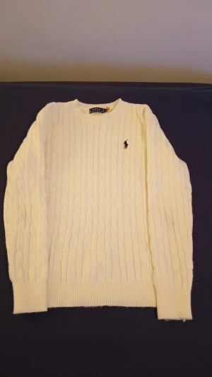Vit Polo Ralph Lauren stickad tröja  - Säljer en knappt använd (använd bara 3 gånger) vit stickad tröja från Polo Ralph Lauren med klassisk rund hals och en mörkblå broderad logga på bröstet.Tröjan har långa ärmar och ribbade muddar vid ärmslut och nederkant. Perfekt för dig som gillar stilren och tidlös design.