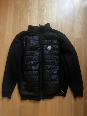 Svart pufferjacka från Moncler i storlek M - Svart pufferjacka från Moncler i storlek M. Jackan har quiltad framsida med glansigt material och två dragkedjeförsedda fickor. Hög krage och Moncler-logga på bröstet. Ärmar och rygg i mjukare tyg för extra komfort. Perfekt för kyliga dagar. Priset kan diskuteras