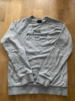 Grå sweatshirt från Peak Performance - Säljer en stilren grå sweatshirt från Peak Performance med svart broderad logga på bröstet. Tröjan har rund hals, långa ärmar och ribbade muddar vid ärmslut och nederkant. Perfekt för en avslappnad och sportig look.