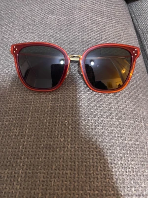 Röda solglasögon från Céline - Snygga solglasögon från Céline med röd, fyrkantig båge och tunna guldfärgade skalmar. Glasen är mörka och bågen har små vita detaljer vid hörnen. Tillverkade i Italien, perfekt för dig som vill ha en cool och stilren look.   Nypris:3000kr