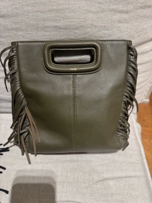 Olivgrön fransad handväska från Maje - Cool olivgrön handväska från Maje i skinn med fransar längs sidorna och inbyggt handtag. Väskan har en stilren, fyrkantig form och diskret logga framtill. Perfekt för dig som vill sticka ut med en trendig accessoar.