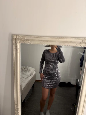 Glittrig silverpaljettklänning - Säljer en glittrig silverfärgad kort klänning med långa ärmar och öppen rygg. Klänningen är täckt av paljetter och har en tight passform som framhäver figuren. Perfekt för dig som vill sticka ut och glänsa på festen!