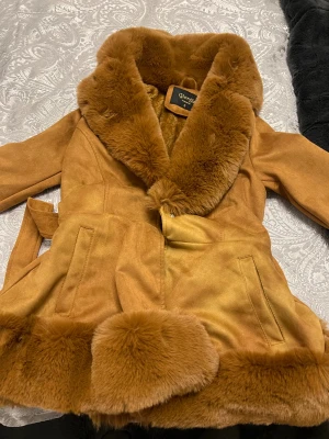 Brun pälsjacka från Giorgia Fashion - Säljer en brun pälsjacka från Giorgia Fashion i storlek S. Jackan har fluffig fuskpäls på krage, insida och nederkant, samt ett bälte i midjan för snygg passform. Perfekt för dig som vill ha en varm och trendig look under höst och vinter.