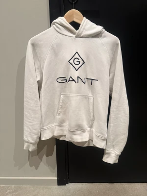 Vit hoodie från GANT med logga - Vit hoodie från GANT med stor broderad logga på bröstet. Klassisk känguruficka framtill, huva och långa ärmar. Mjuk och skön bomullskänsla, perfekt för chill dagar. Enkel och stilren design som funkar till det mesta.