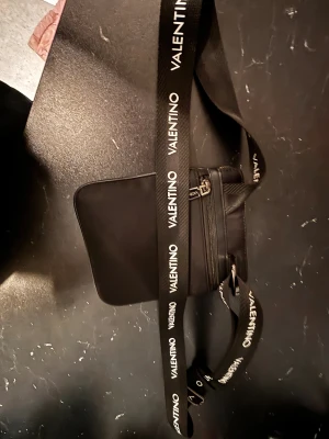 Svart axelväska från Valentino - Snygg svart axelväska från Valentino med justerbart band där loggan är tryckt i vitt. Väskan har en dragkedja framtill och är tillverkad i ett slitstarkt syntetmaterial. Perfekt storlek för mobil, plånbok och andra småsaker.