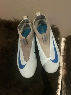 Nike fotbollsskor i vitt och blått - Säljer ett par Nike fotbollsskor med stilren design i vitt och blått. Skorna har en slät ovandel med stor blå Swoosh, grått skaft och mönstrad yttersida. Utrustade med dobbar för gräsplan och tillverkade i syntetmaterial för lätt känsla på foten.