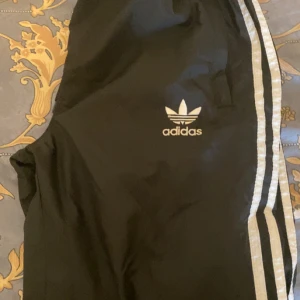 Svarta Adidas byxor med vita ränder - Svarta Adidas byxor med klassiska vita ränder längs sidorna och Adidas Trefoil-logga på benet. Byxorna har resår i midjan och är tillverkade i ett lätt, syntetiskt material som passar perfekt för träning eller chill. Snygg och sportig look som aldrig går ur tiden.