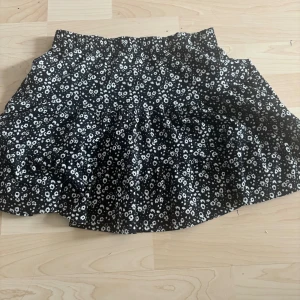 Svart blommig kortkjol - Supersöt svart kortkjol med vita små blommor över hela tyget. Kjolen har en lätt utställd form och resår i midjan för extra komfort. Perfekt att styla med en enkel topp och sneakers för en chill look.