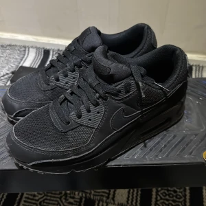 Nike Air Max 90 Triple Black sneakers - Säljer ett par helsvarta Nike Air Max 90 sneakers med klassisk siluett. Skorna har ovandel i mesh och skinn, synlig Air Max-dämpning i sulan och tydliga Nike-loggor på häl och tunga. Perfekta för dig som gillar en clean och sportig look.använd typ 3-4 gånger 
